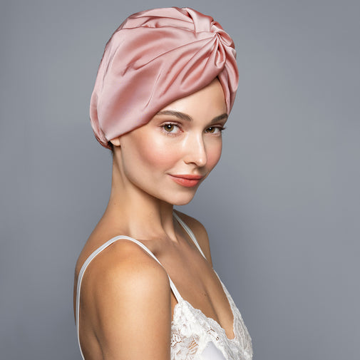 A woman wearing a satin turban-style hair wrap in dusty pink, photographed against a plain gray studio background.