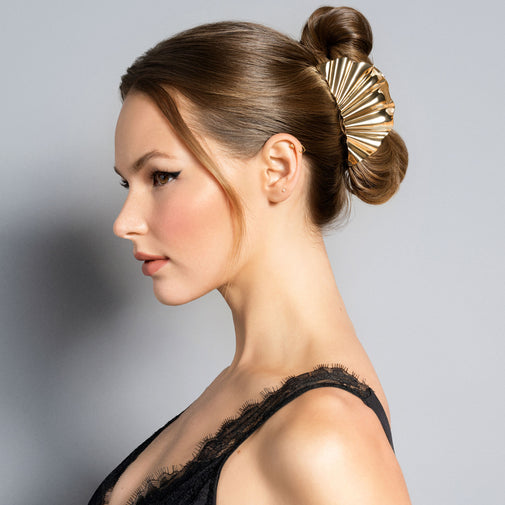 A woman wearing a metallic gold, fan-shaped hair claw clip holding her hair in a bun, set against a solid gray studio background.