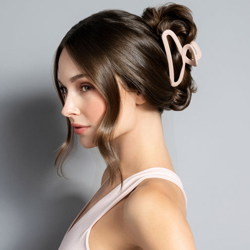 A woman in profile wearing a pale blush pink claw clip that holds her brown hair in a French twist, against a plain light gray studio background.