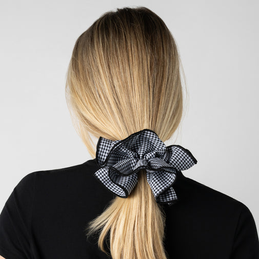 A woman shown from the back with long blonde hair tied in a low ponytail with a large black-and-white gingham bow scrunchie, wearing a black top against a light gray background.