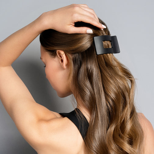 A woman with long, wavy light brown hair wearing a matte black square-shaped hair claw clip, shown from a side-back angle against a solid gray background.