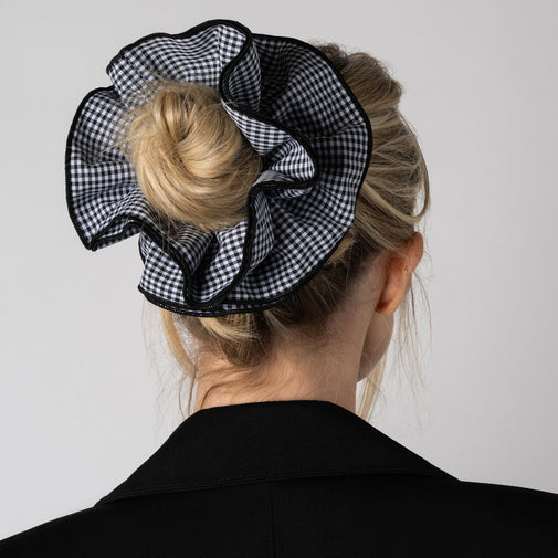 A black and white gingham ruffled hair scrunchie worn on a blonde woman's bun, shown from the back against a light gray background.