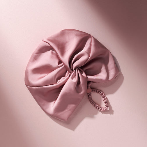 A dusty pink satin hair bonnet with a matching fabric elastic band, styled on a soft pink background with artistic diagonal shadows.
