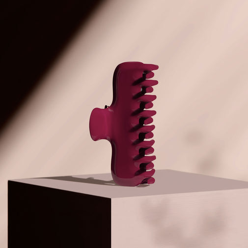 A single hair claw clip in a dark red color, standing upright on a white display block against a beige wall with dramatic diagonal shadows.