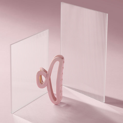 A pink silicone hair claw clip, displayed in front of two translucent white ribbed acrylic panels on a soft pink background.