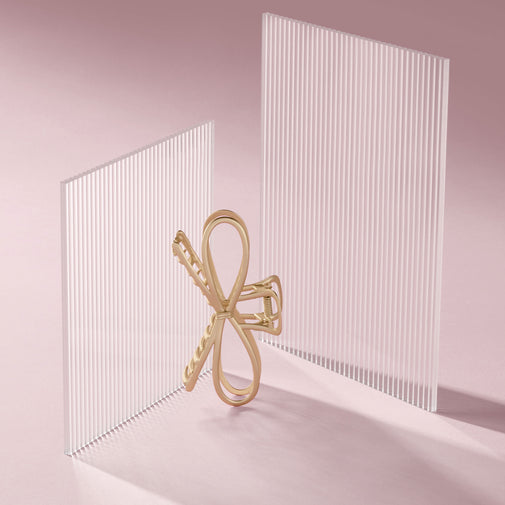 A gold bow shaped hair claw clip stands between two translucent white ribbed panels on a soft pink studio background.