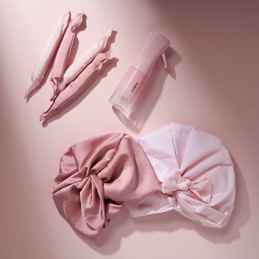 Pink hair styling accessories arranged on a soft pink background, including curl define rollers, a spray bottle, a shower cap and a hair bonnet in dusty pink and pale pink.