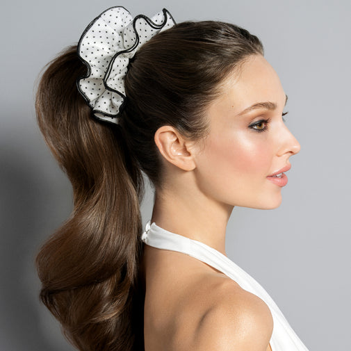 A woman in profile wearing a white ruffled scrunchie with black polka dots and black trim, holding her brown hair in a ponytail against a solid gray background.