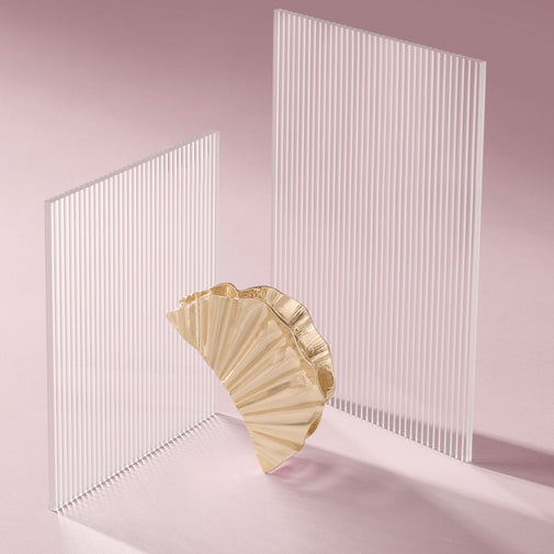 A gold pleated fan-shaped claw clip stands in front of two translucent white slatted panels on a soft pink studio background.