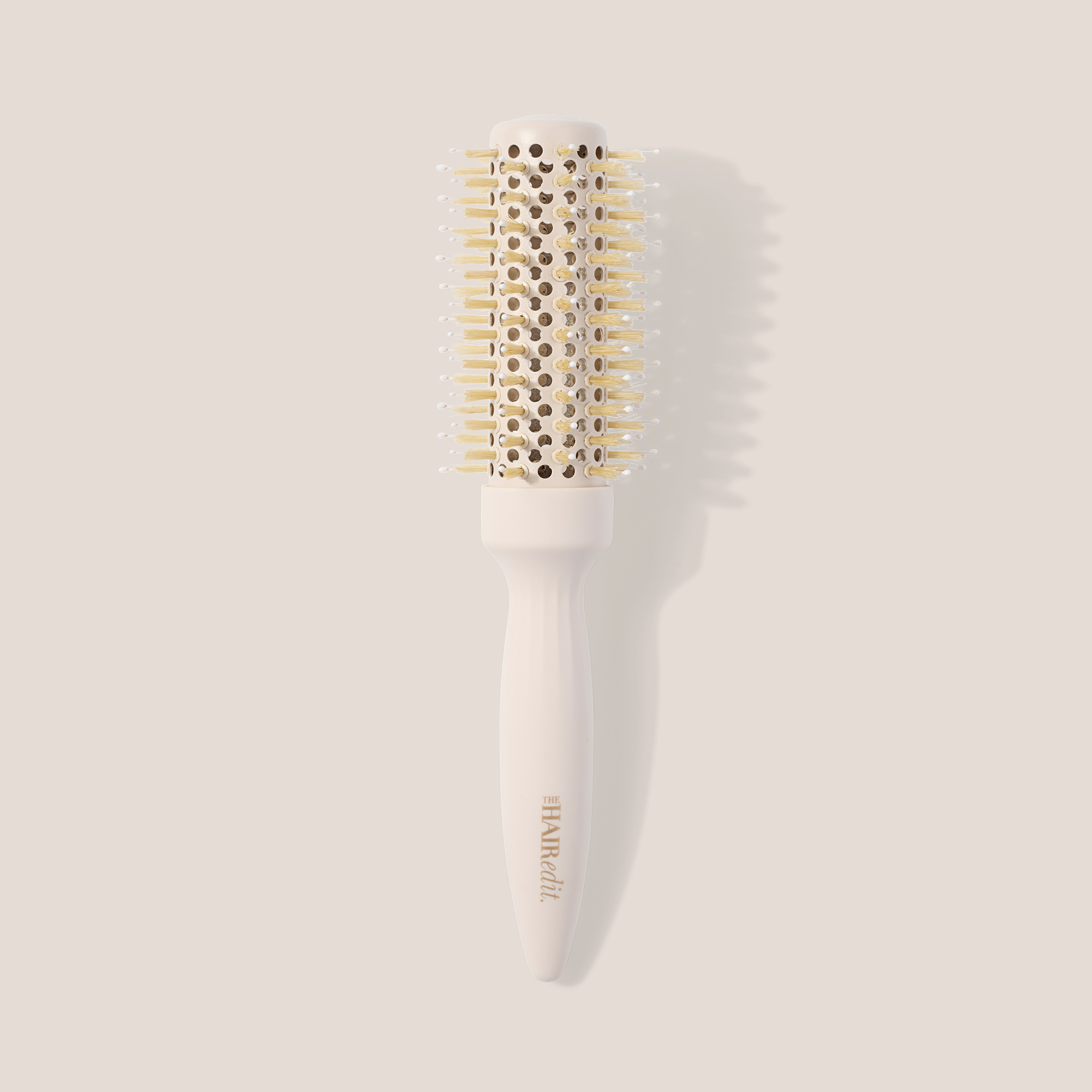 A round hairbrush with a white handle and barrel, featuring light gold bristles and a gold-embossed brand name, set against a solid beige background with a soft shadow.1