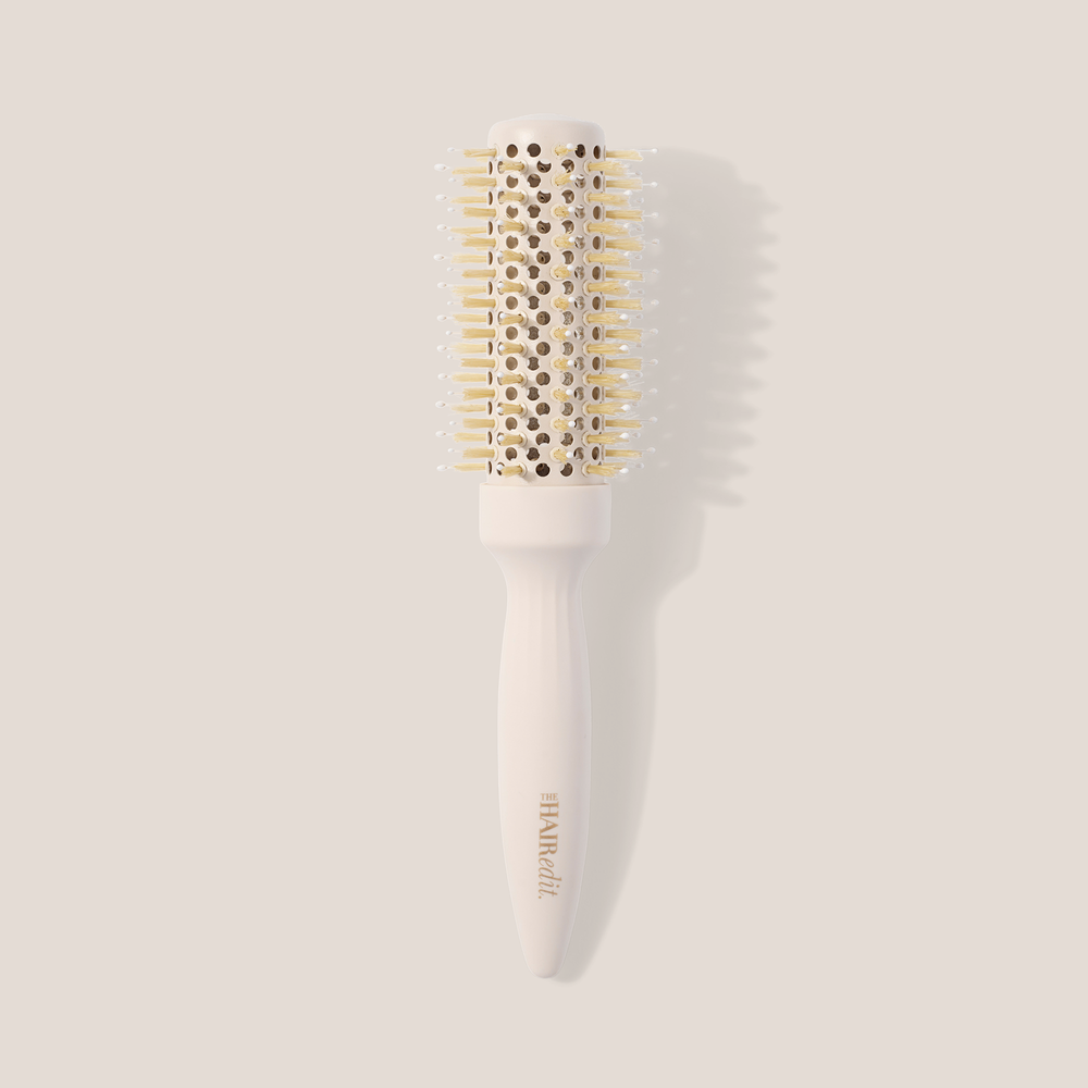 A round hairbrush with a white handle and barrel, featuring light gold bristles and a gold-embossed brand name, set against a solid beige background with a soft shadow.1