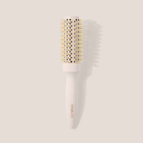 A round hairbrush with a white handle and barrel, featuring light gold bristles and a gold-embossed brand name, set against a solid beige background with a soft shadow.