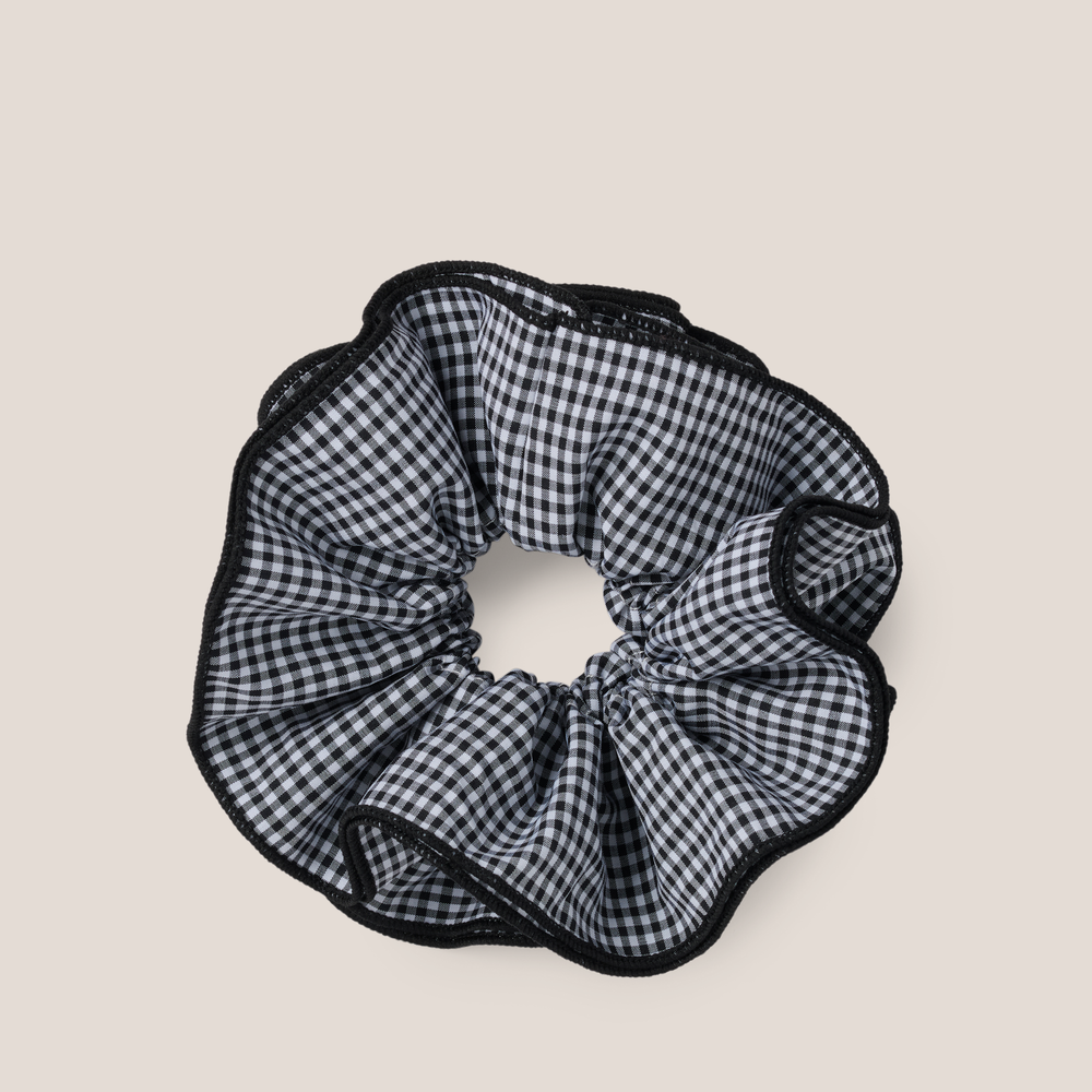 A black and white gingham check scrunchie with black trim, shown on a solid light beige background.1
