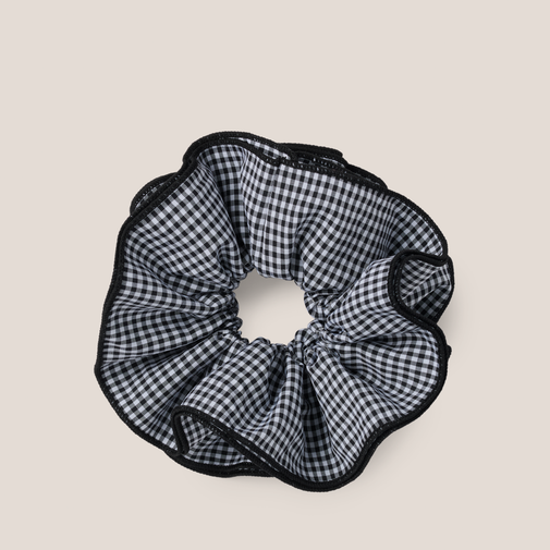 A black and white gingham check scrunchie with black trim, shown on a solid light beige background.