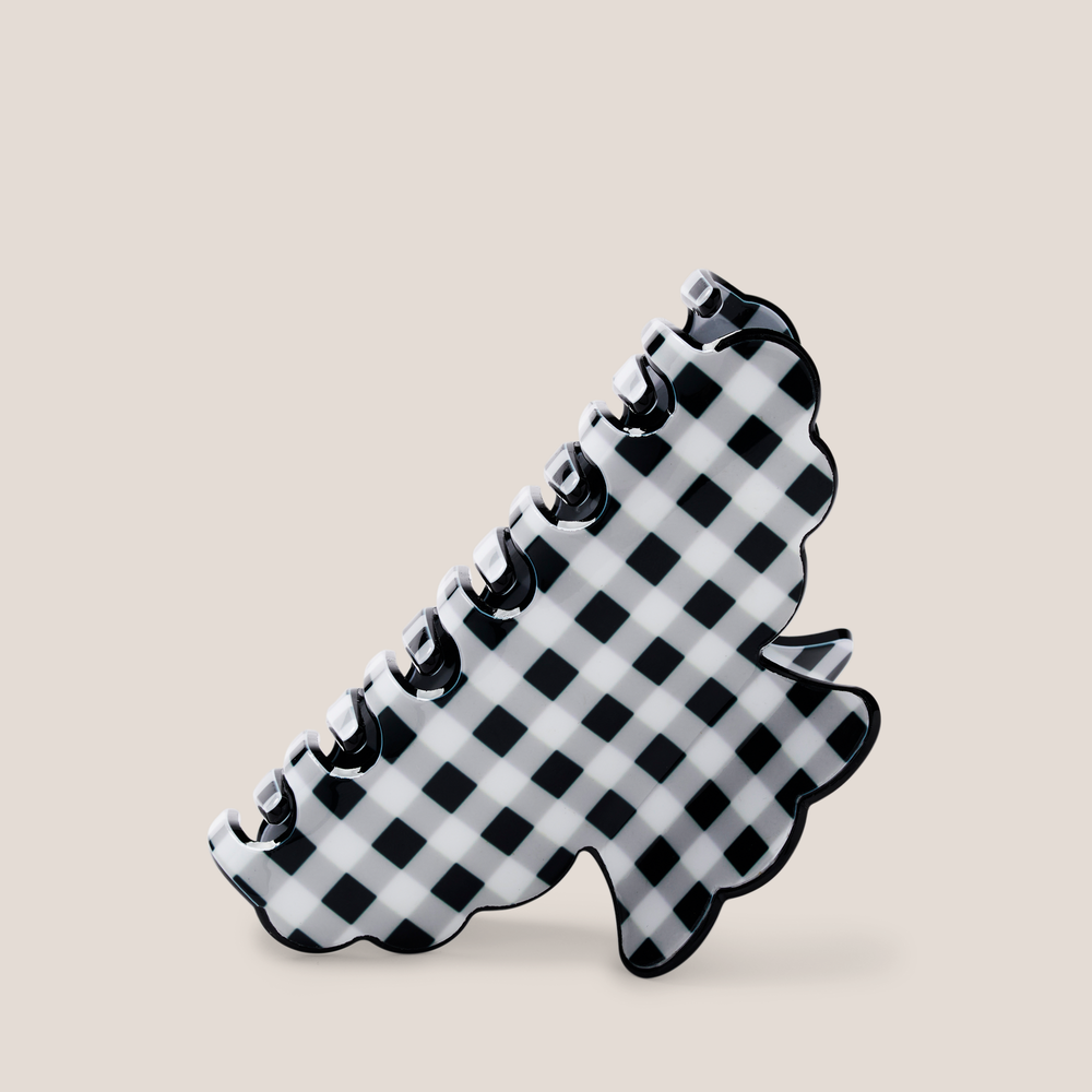 A black and white checkered claw hair clip with a scalloped edge, shown on a solid light beige background.1