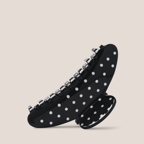 A black claw hair clip with white polka dots and silver metal spring teeth, shown on a solid light beige background.