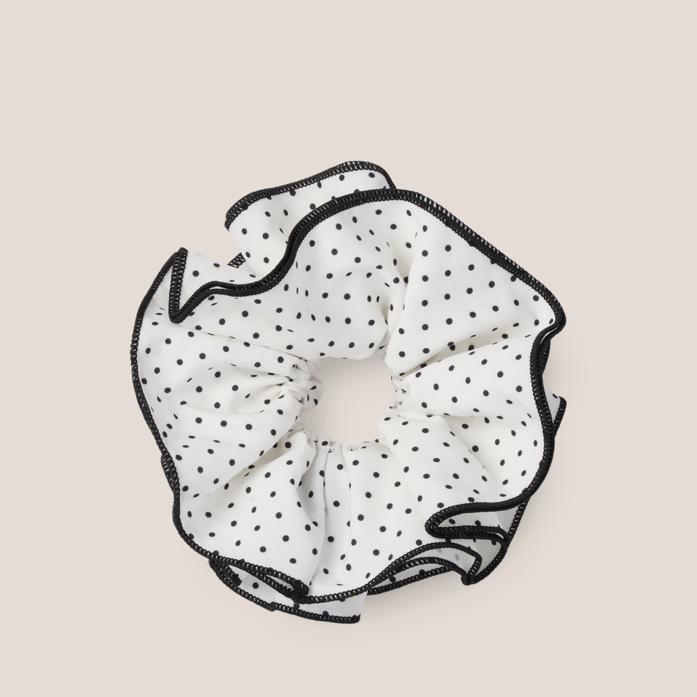 A white ruffled scrunchie with black polka dots and black stitched trim, shown flat against a light beige background.1