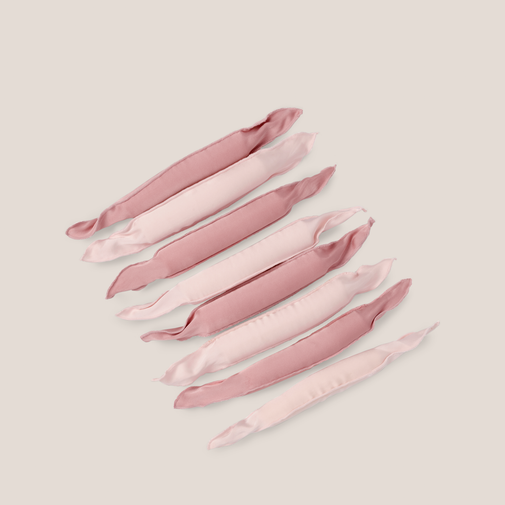 Six satin pillow rollers in alternating dusty pink and pale blush pink, arranged diagonally on a solid light beige background.