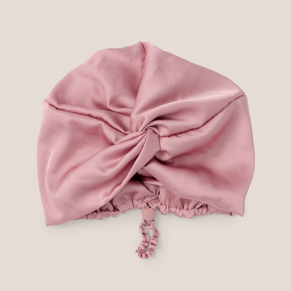 A dusty pink satin bonnet with a gathered twist at the top and a matching drawstring tie, displayed on a neutral light beige background.1