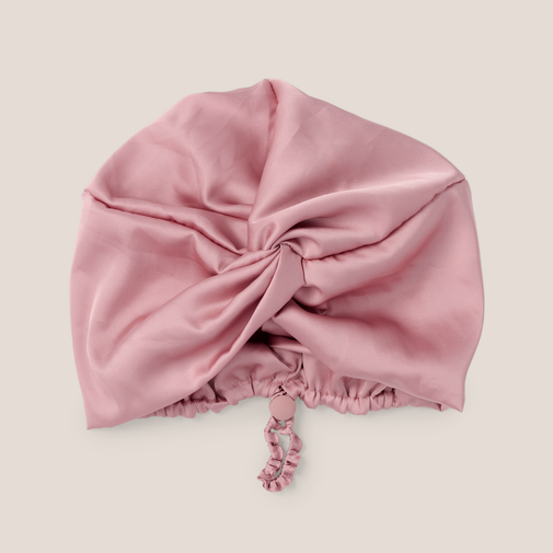 A dusty pink satin bonnet with a gathered twist at the top and a matching drawstring tie, displayed on a neutral light beige background.