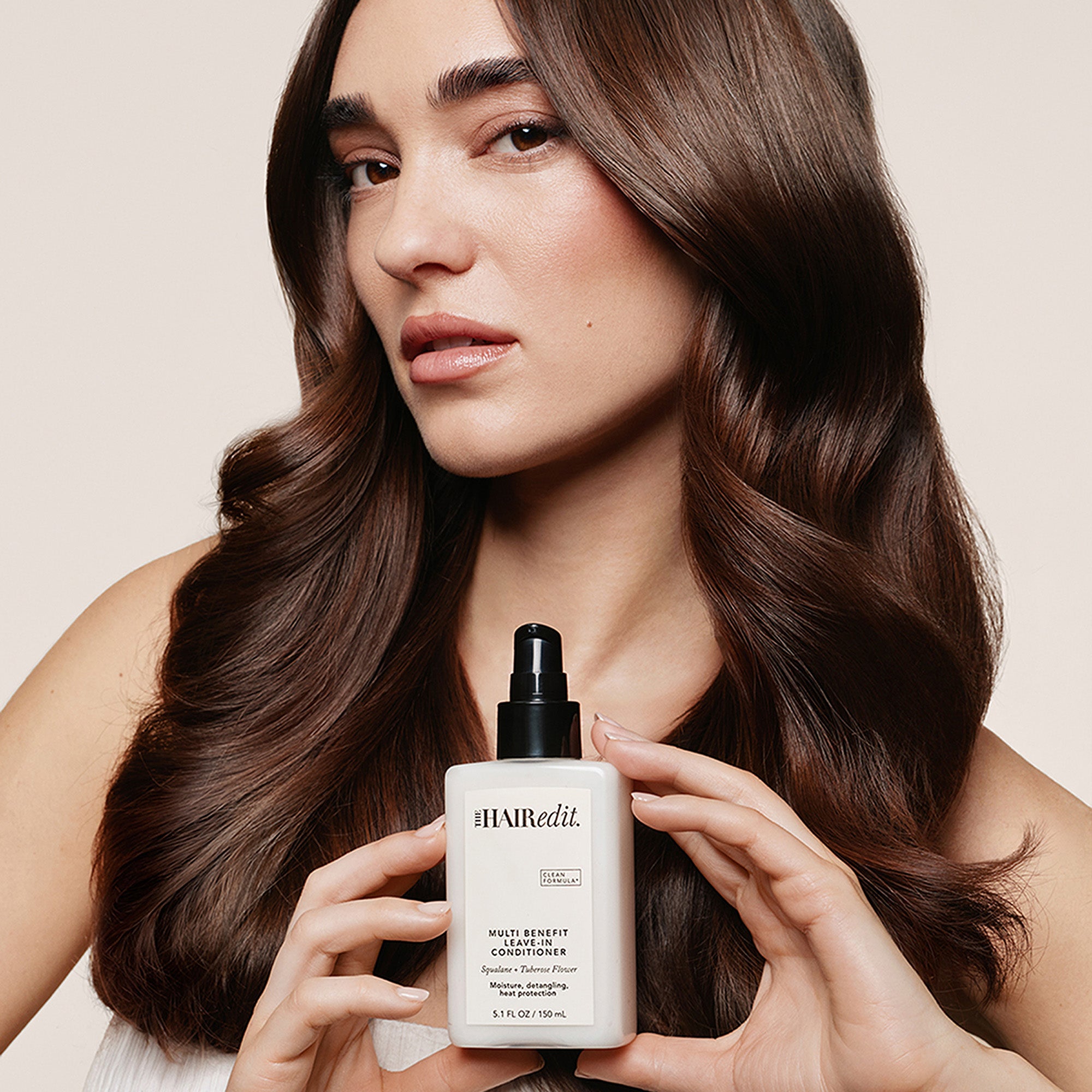 A woman with long, wavy brown hair holds a white bottle of The Hair Edit Multi-Benefit Conditioner against a warm beige background.10