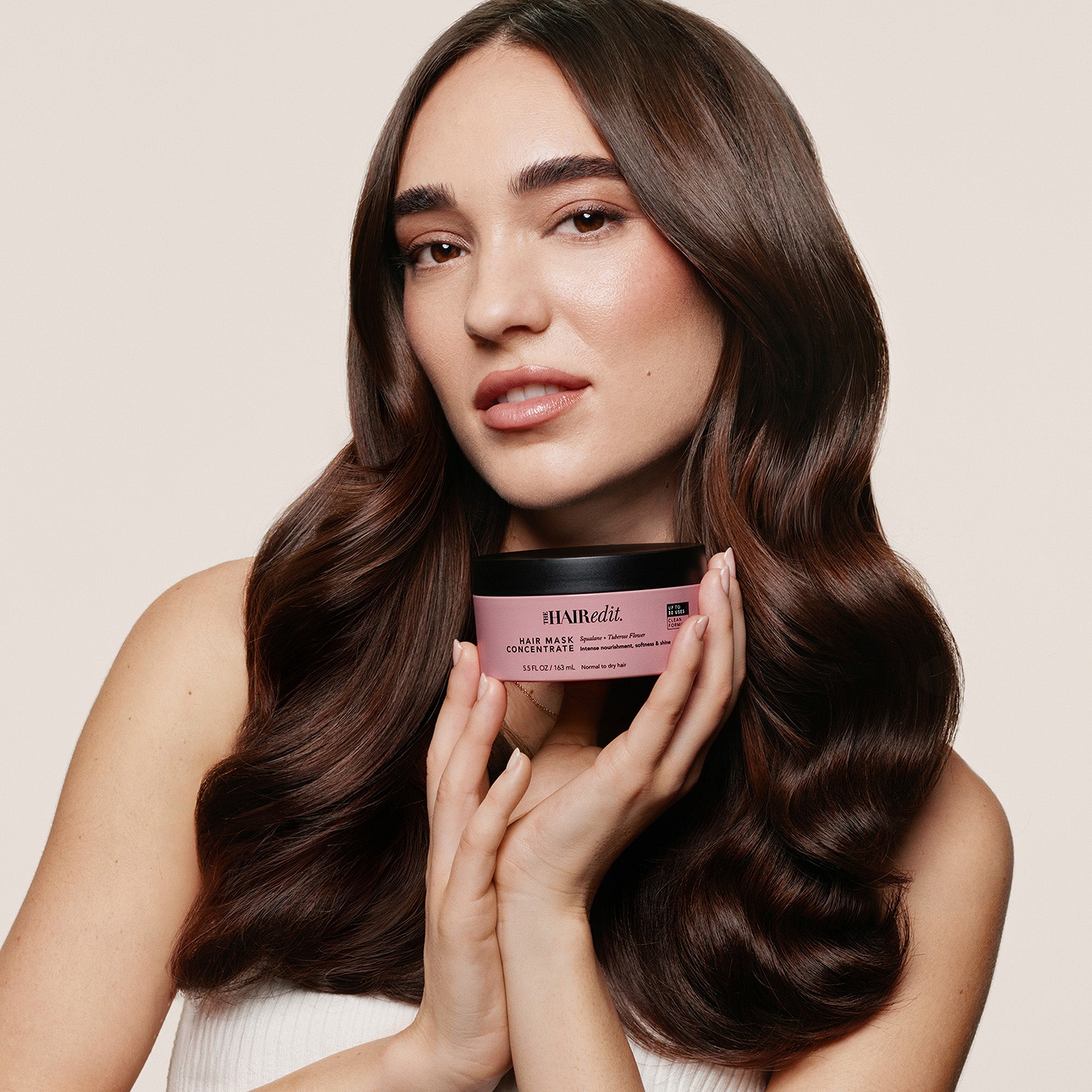 A woman with long, wavy brown hair holds a pink jar of The Hair Edit Hair Mask Concentrate against a warm beige background.11