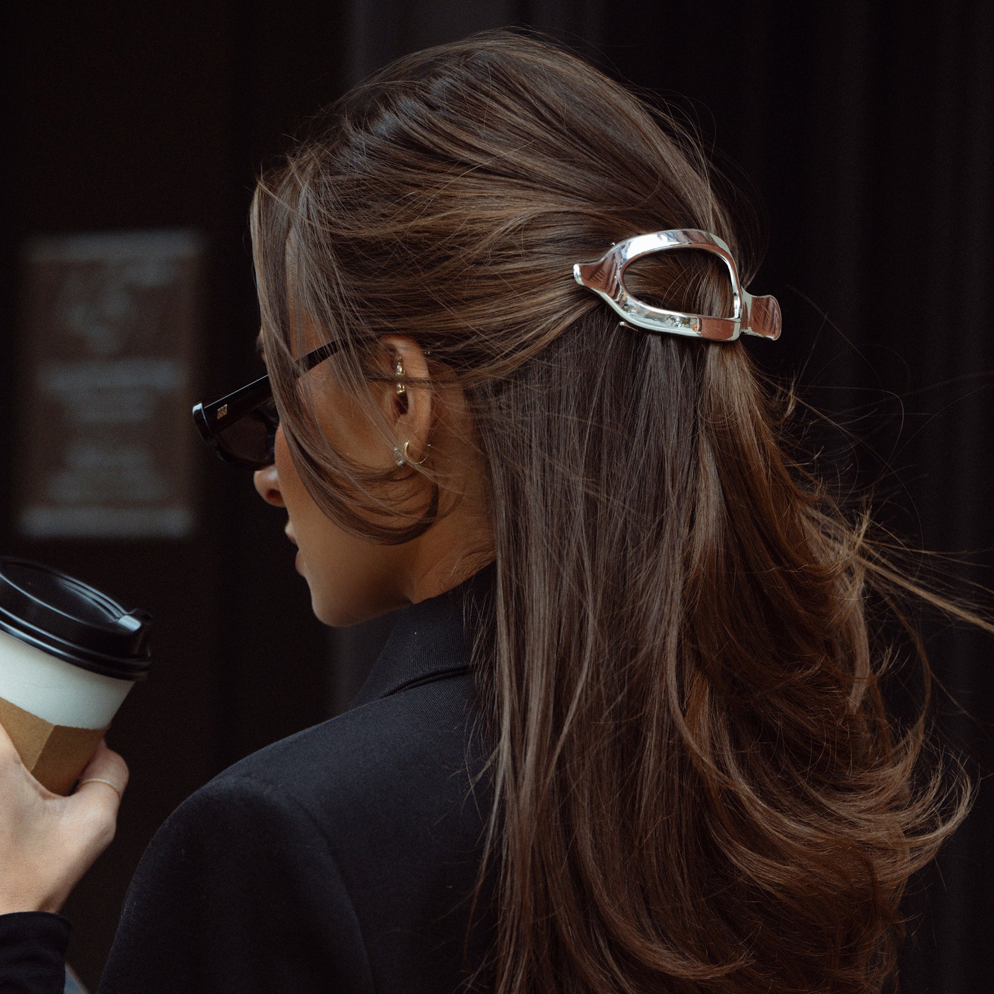 A woman in side profile wearing a metallic silver flat lay claw-style hair clip that holds back a brown highlighted ponytail. She is wearing black sunglasses and a black blazer, holding a takeaway coffee cup against a dark background.3