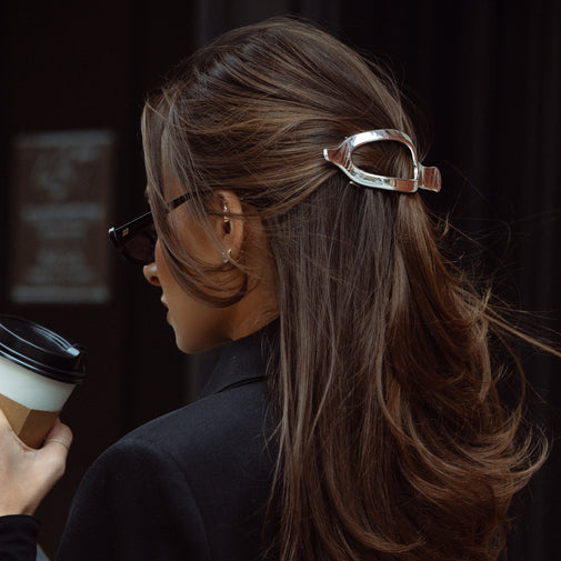 A woman in side profile wearing a metallic silver flat lay claw-style hair clip that holds back a brown highlighted ponytail. She is wearing black sunglasses and a black blazer, holding a takeaway coffee cup against a dark background.