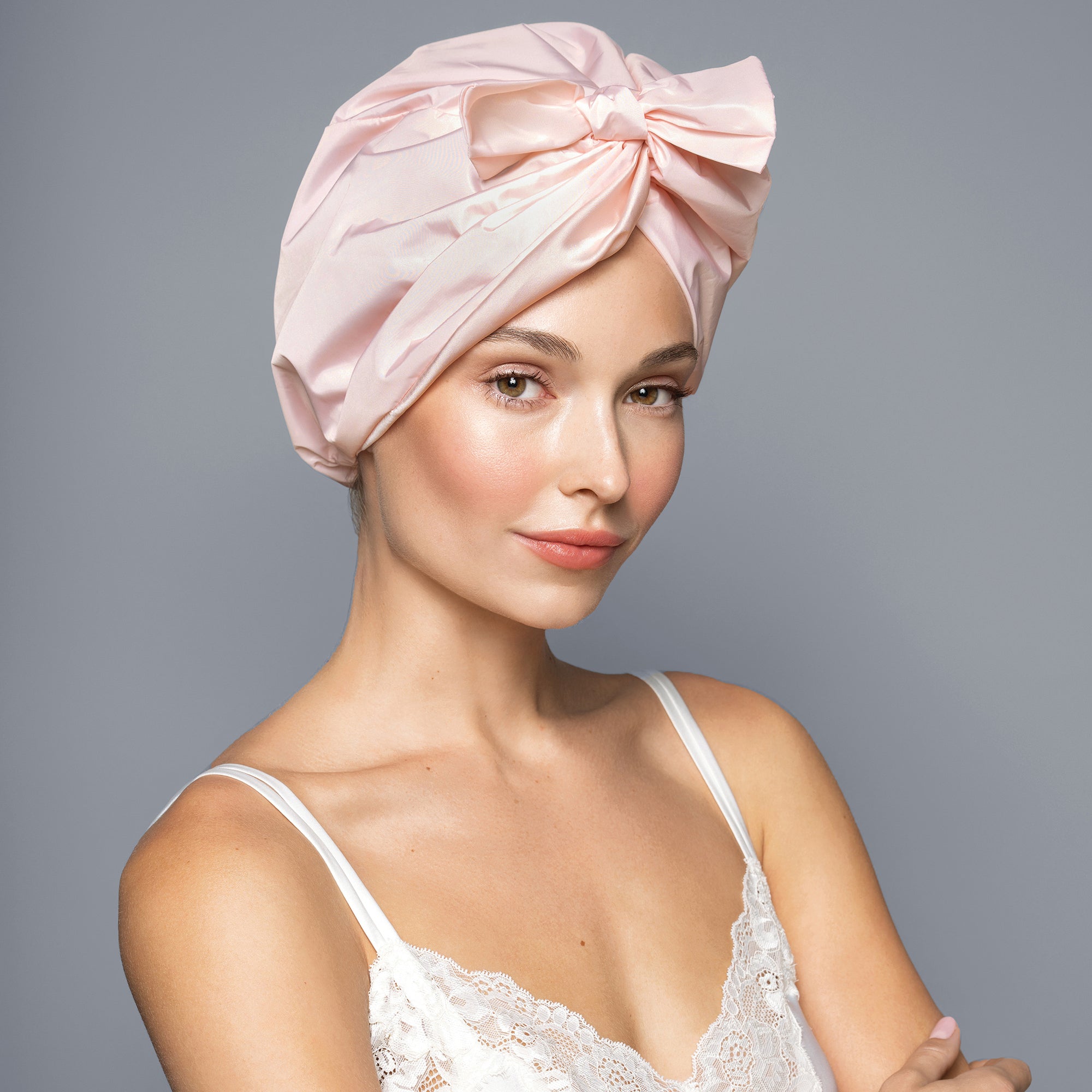 A woman wearing a light pink satin turban headwrap with a large bow, styled on her head against a solid gray studio background.3