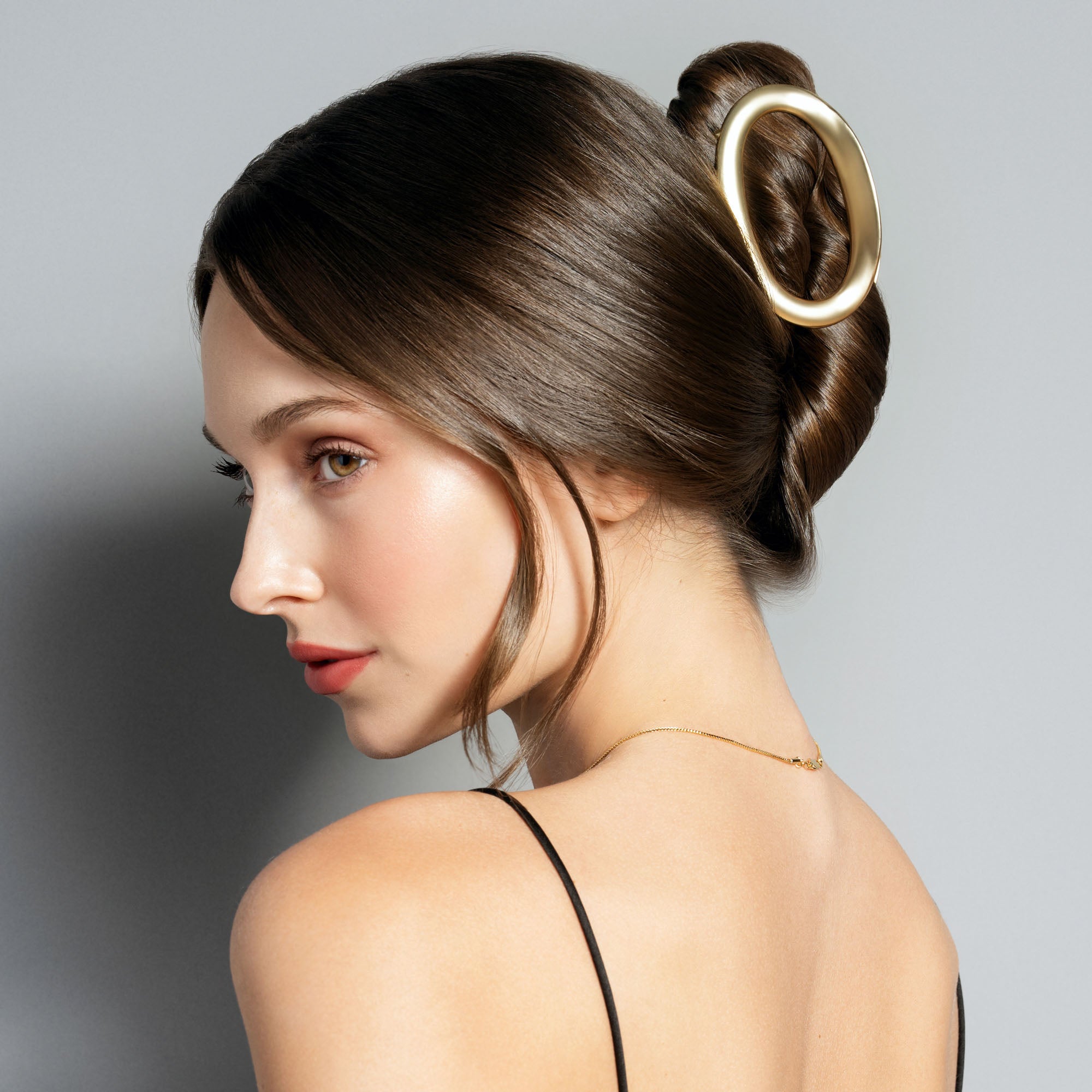 A woman shown in side profile with her hair styled in a low bun, wearing a large oval gold hair claw clip. The background is a clean, neutral gray studio backdrop.3