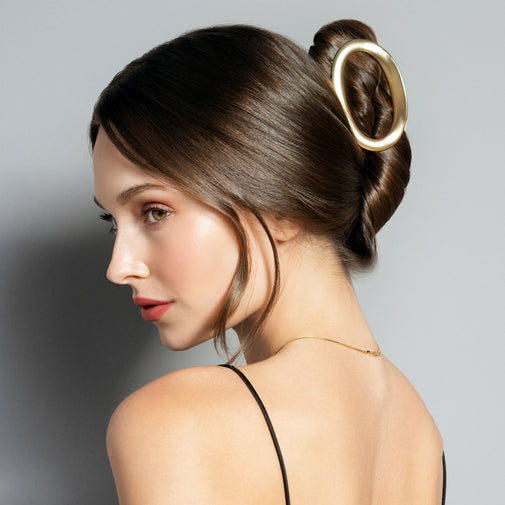 A woman shown in side profile with her hair styled in a low bun, wearing a large oval gold hair claw clip. The background is a clean, neutral gray studio backdrop.