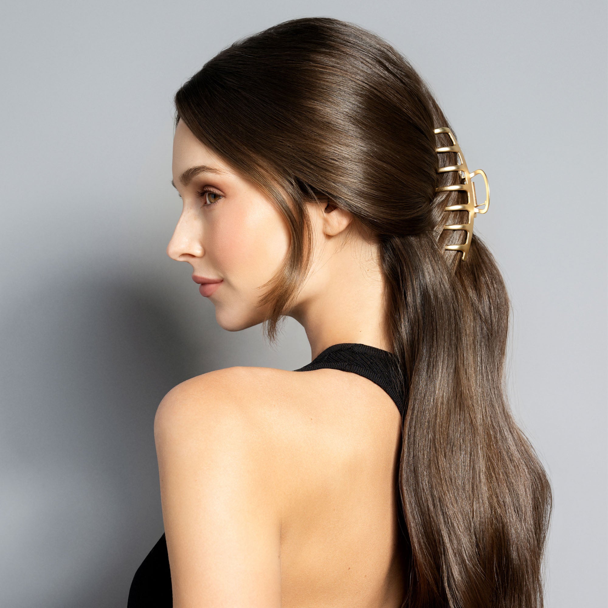 A woman with long, straight brown hair is shown in profile wearing a gold claw-style hair clip to secure a low ponytail. She is wearing a black halter top against a solid gray studio background.3
