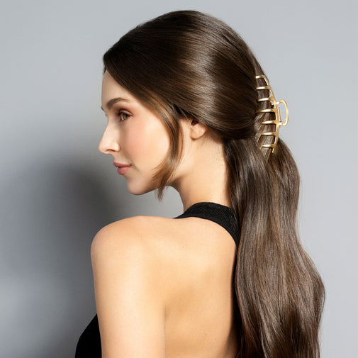 A woman with long, straight brown hair is shown in profile wearing a gold claw-style hair clip to secure a low ponytail. She is wearing a black halter top against a solid gray studio background.