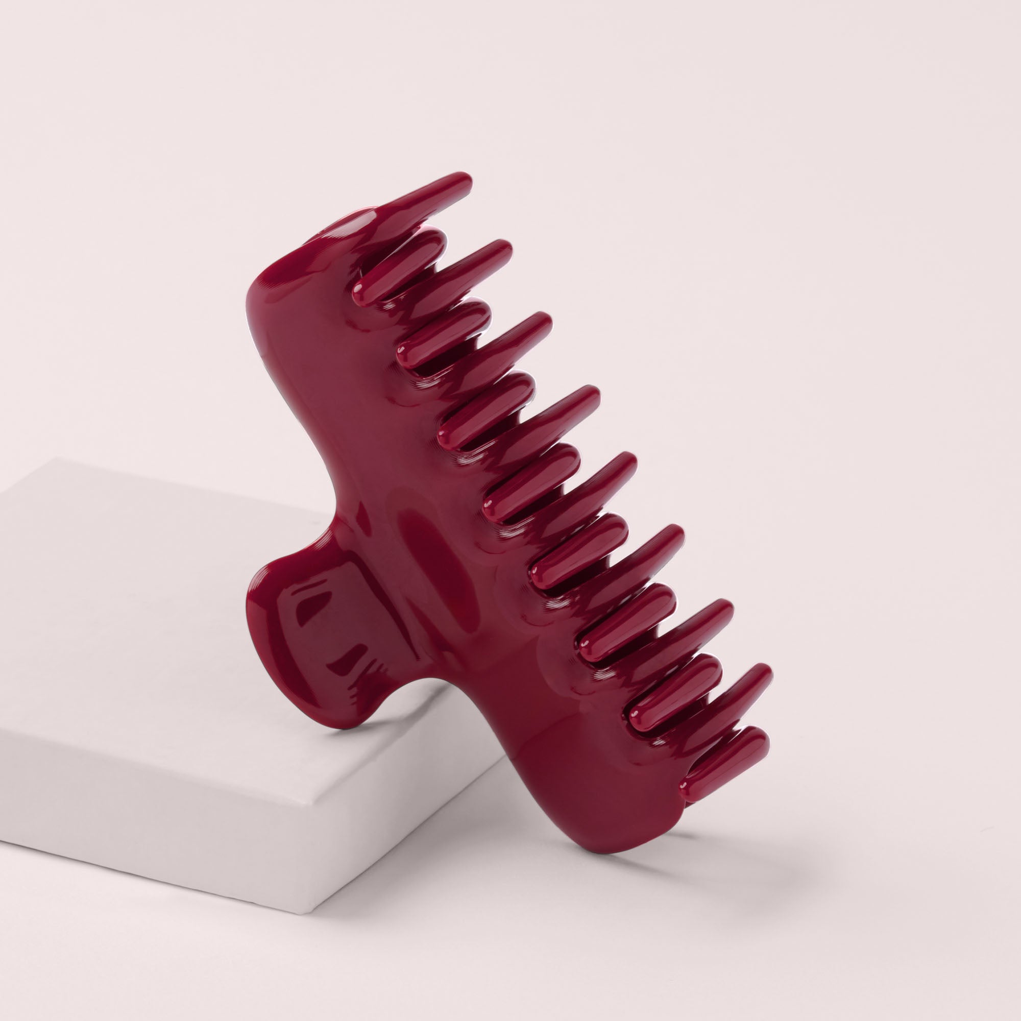 A glossy dark red hair claw comb resting on a pink display block against a light pink background.4