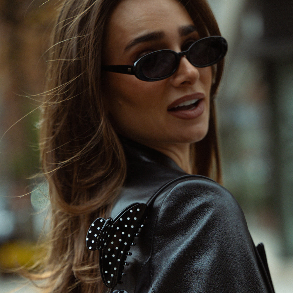 A close-up lifestyle shot of a woman wearing black oval sunglasses and a black leather jacket with a polka-dot bow detail on the shoulder, set against a blurred outdoor background.4
