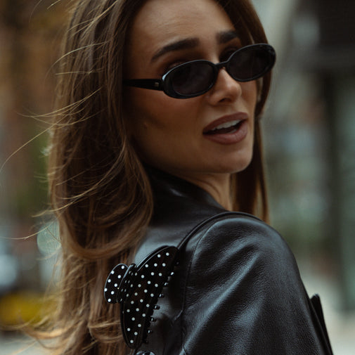 A close-up lifestyle shot of a woman wearing black oval sunglasses and a black leather jacket with a polka-dot bow detail on the shoulder, set against a blurred outdoor background.