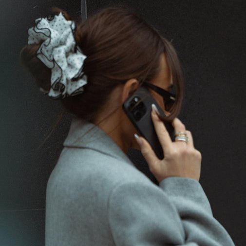 A woman in profile wearing a large white hair scrunchie with black polka dots and black trim, holding a black smartphone to her ear. She is wearing a light gray coat against a dark gray background.