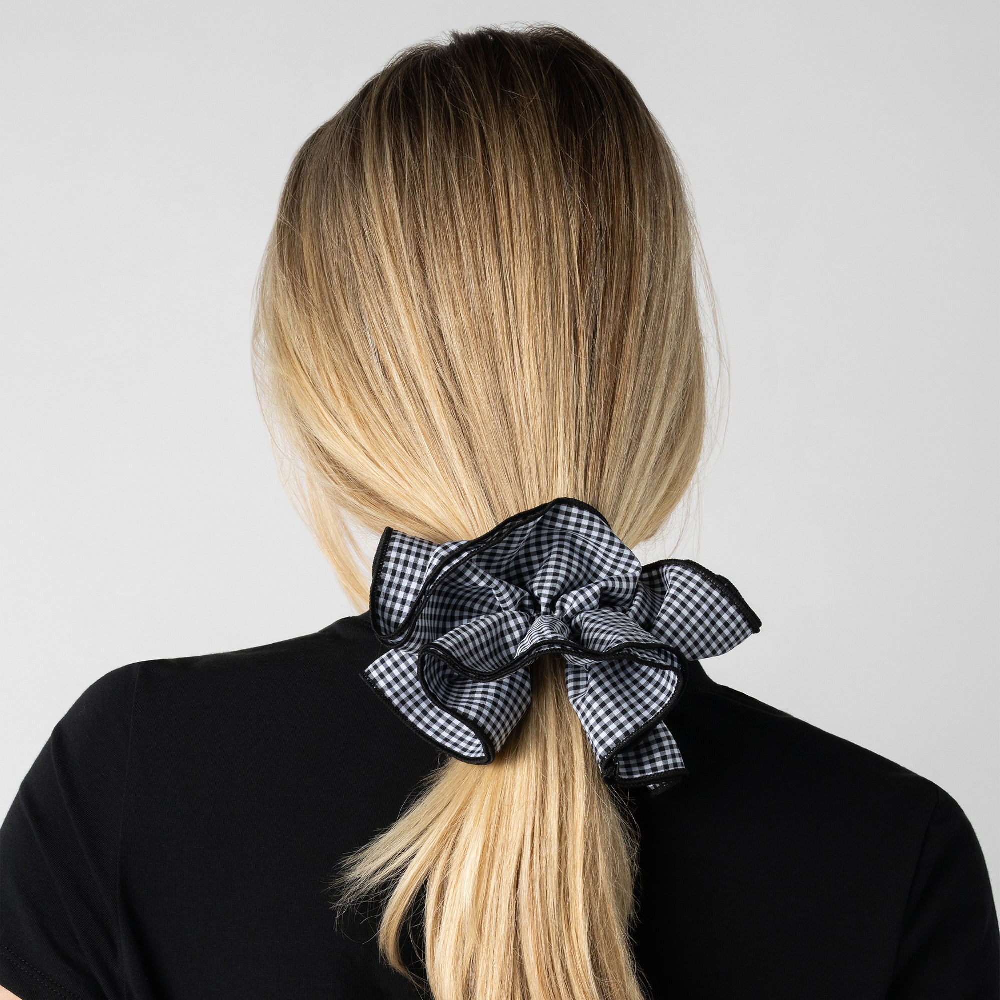 A woman shown from the back with long blonde hair tied in a low ponytail with a large black-and-white gingham bow scrunchie, wearing a black top against a light gray background.6