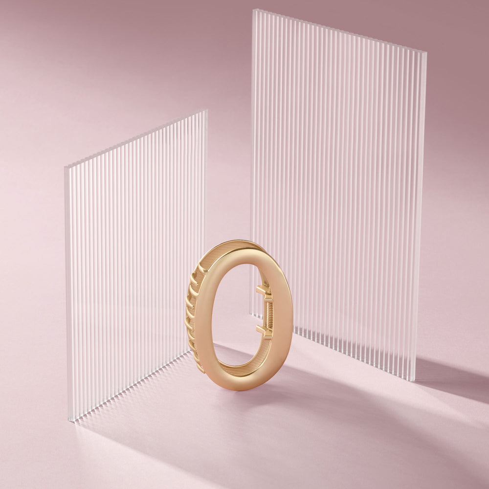 A gold-toned circular ring claw clip, displayed on a soft pink studio surface with two translucent vertical slatted panels behind it.4
