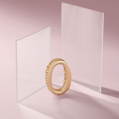 A gold-toned circular ring claw clip, displayed on a soft pink studio surface with two translucent vertical slatted panels behind it.
