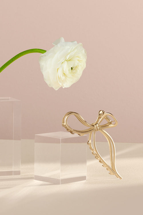The Hair Edit Gold Gilded Bow Claw Clip in a lifestyle setting with a white flower 