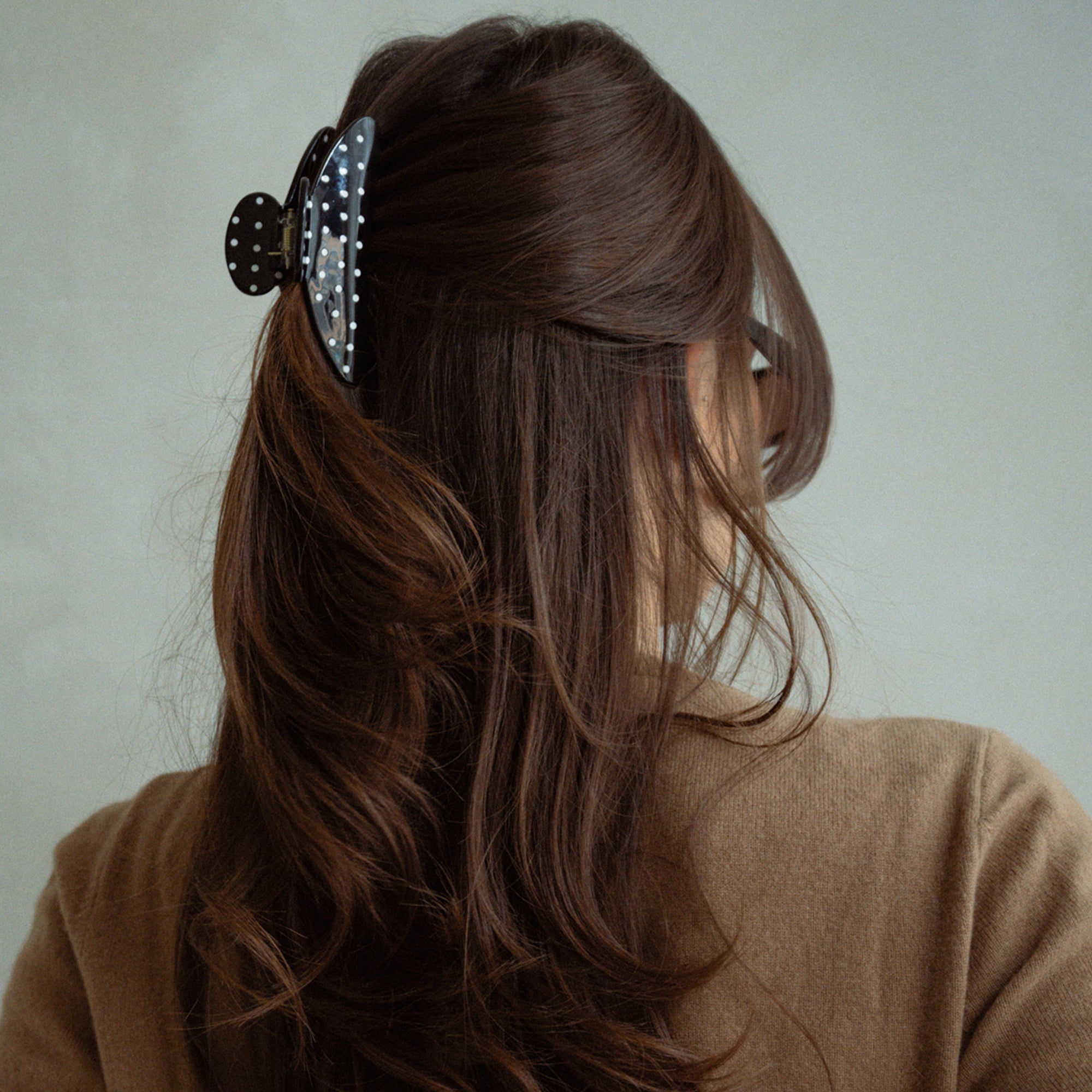A woman shown from the back wearing a large black claw hair clip with white polka dots, holding back her long brown hair. She is wearing a brown knit top against a plain light gray background.5