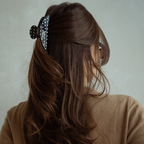 A woman shown from the back wearing a large black claw hair clip with white polka dots, holding back her long brown hair. She is wearing a brown knit top against a plain light gray background.