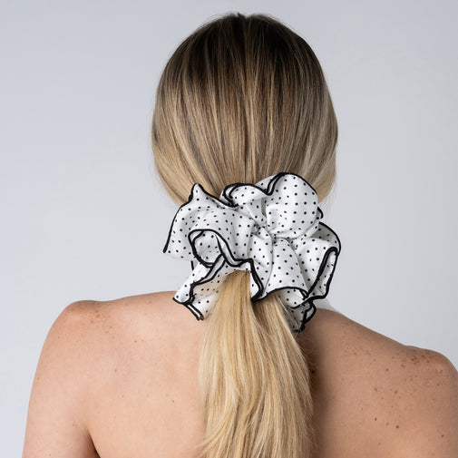 A blonde woman viewed from the back, wearing a large ruffled scrunchie in white fabric with small black polka dots and black trim, holding her hair in a low ponytail against a light gray background.