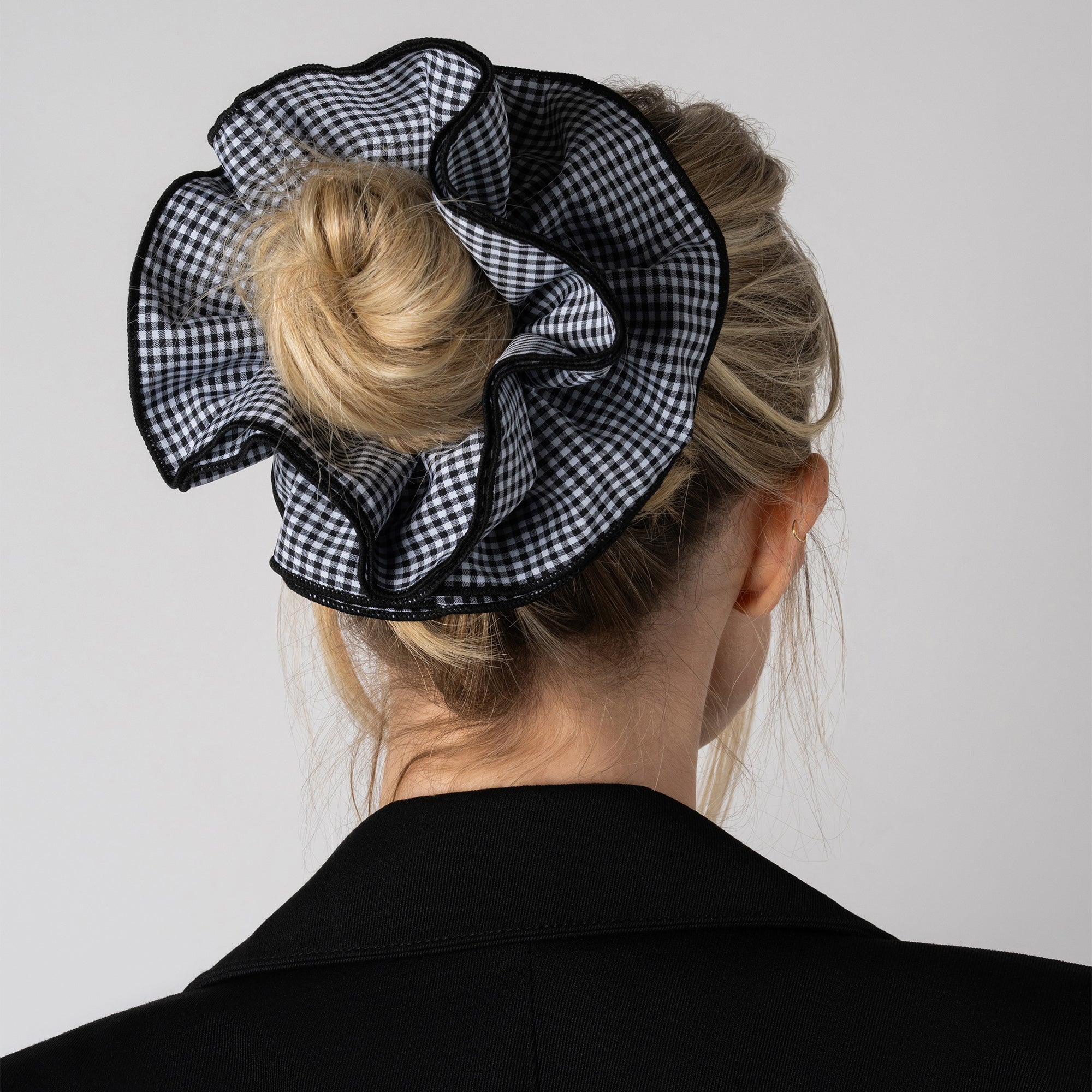 A black and white gingham ruffled hair scrunchie worn on a blonde woman's bun, shown from the back against a light gray background.7