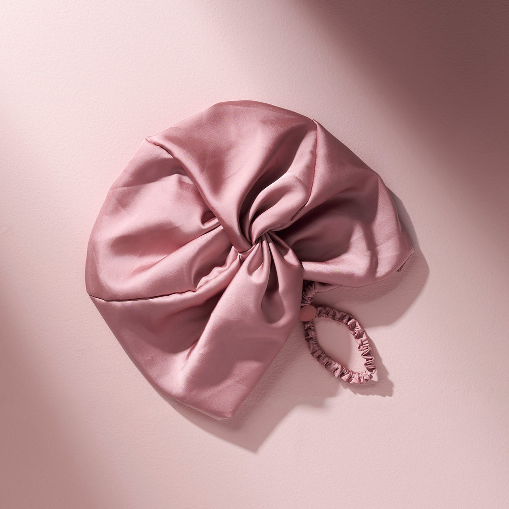 A dusty pink satin hair bonnet with a matching fabric elastic band, styled on a soft pink background with artistic diagonal shadows.5