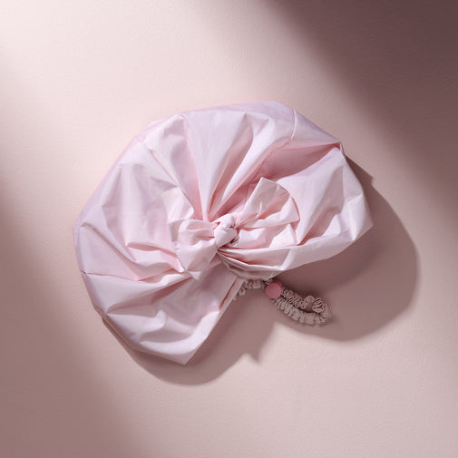 A light pink fabric hair scrunchie with a matching elastic band and a small pink bead accent, styled on a soft pink background with a diagonal shadow.