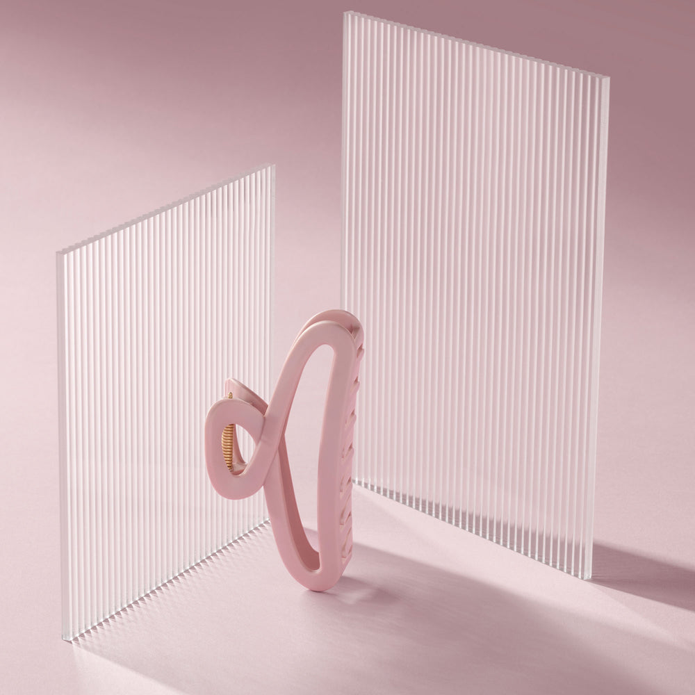 A pink silicone hair claw clip, displayed in front of two translucent white ribbed acrylic panels on a soft pink background.5