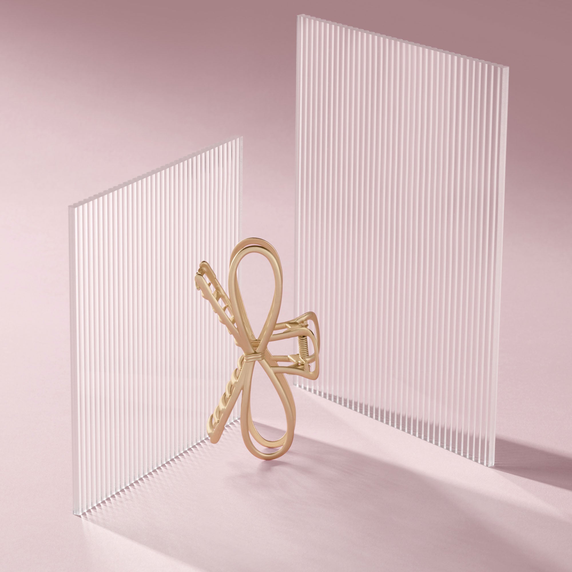 A gold bow shaped hair claw clip stands between two translucent white ribbed panels on a soft pink studio background.5