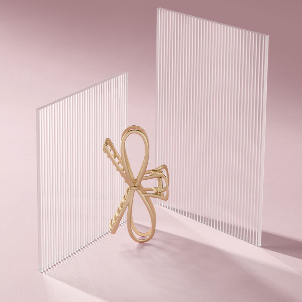 A gold bow shaped hair claw clip stands between two translucent white ribbed panels on a soft pink studio background.5